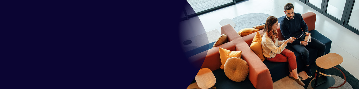 Egy férfi és egy nő ül együtt egy modern irodai lounge térben, egy táblagépet nézve, színes kanapékon, kerek párnákkal és kis oldalasztalokkal.